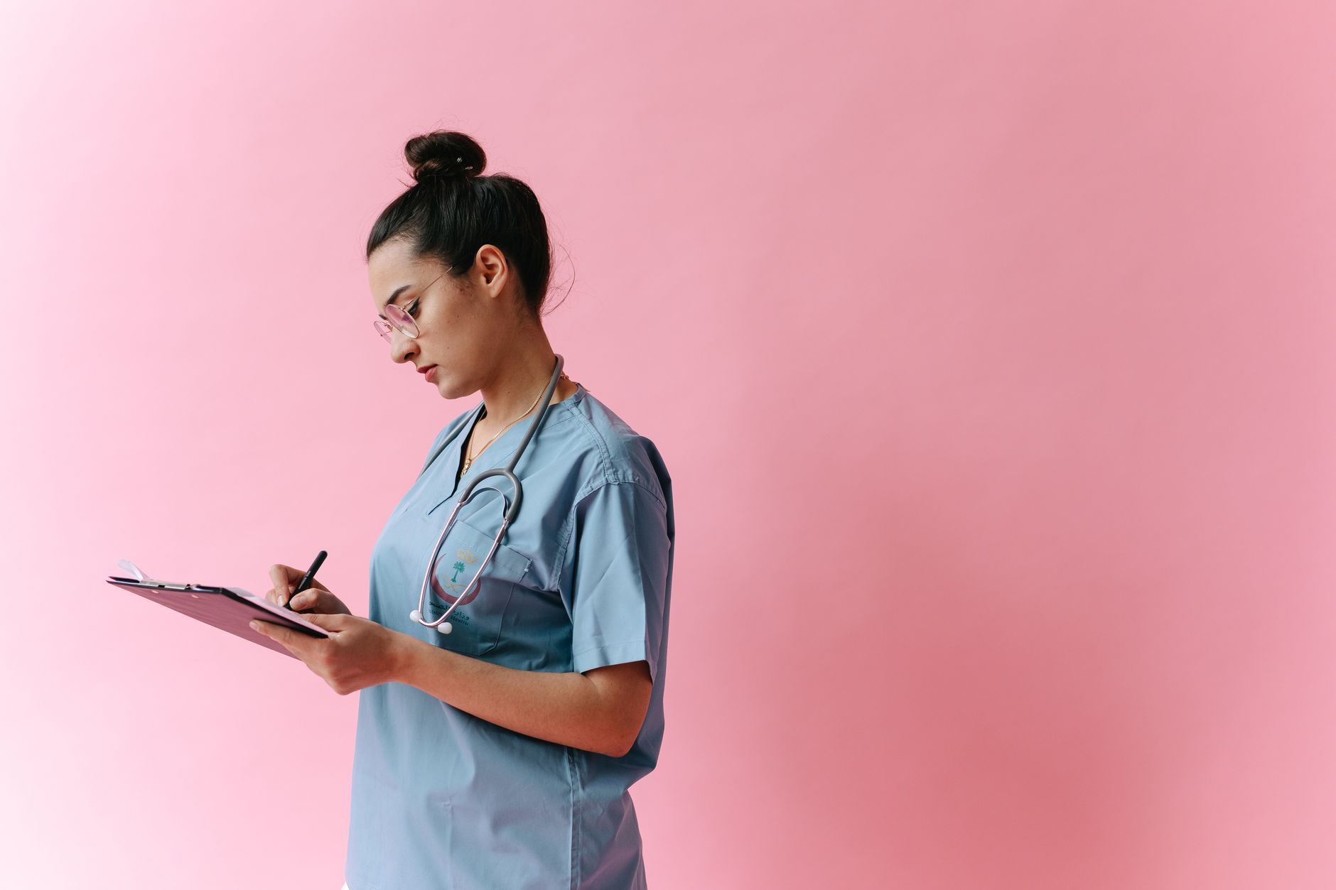 a nurse in scrubs writing notes on a clipboard at a nurse station with a computer monitor nearby